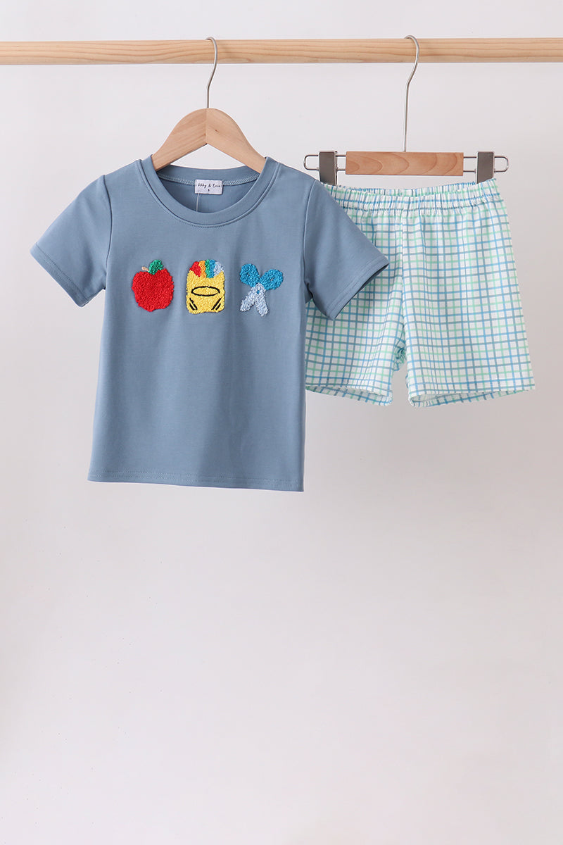 A children's two-piece clothing set hanging on wooden hangers from a light wood rod. The set includes a light blue short-sleeve t-shirt with applique graphics of a red apple, a yellow cup, and a blue bow, paired with light blue and white gingham shorts.