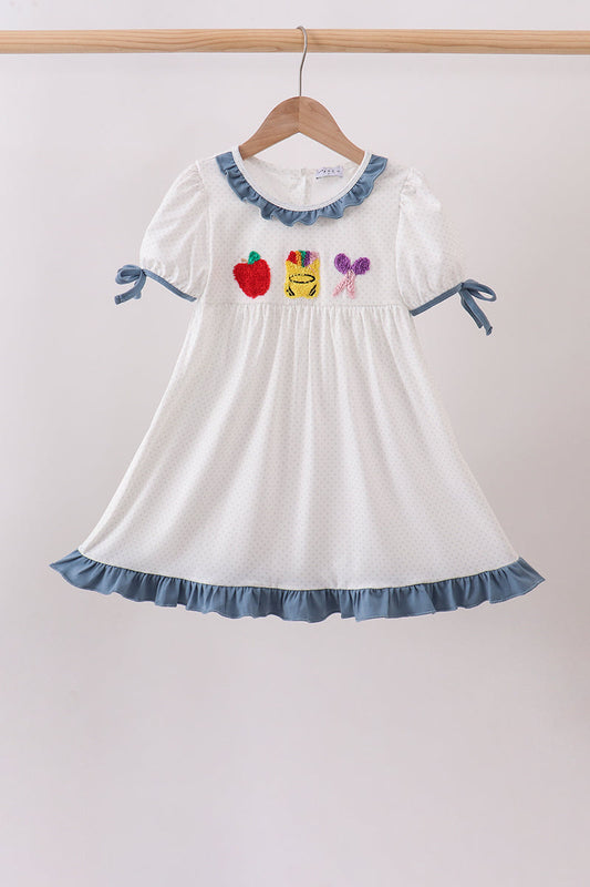A white short-sleeve baby dress with blue ruffle trim, hanging on a wooden hanger against a light beige wall. The dress features three colorful embroidered motifs on the chest: a red apple, a yellow bee, and a purple bow.