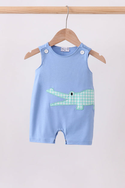 A baby romper in light blue, hanging on a wooden hanger. The romper features a light green and white gingham crocodile applique on the front.