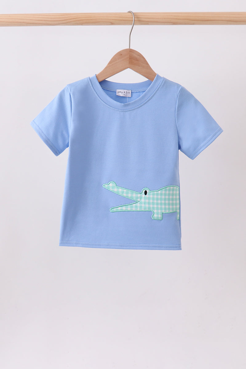 A light blue short-sleeve kids' t-shirt hanging on a wooden hanger against a plain light gray wall. The shirt features a green and white gingham patterned crocodile applique on the front.
