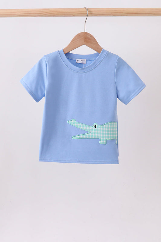 A light blue short-sleeve kids' t-shirt hanging on a wooden hanger against a plain light gray wall. The shirt features a green and white gingham patterned crocodile applique on the front.