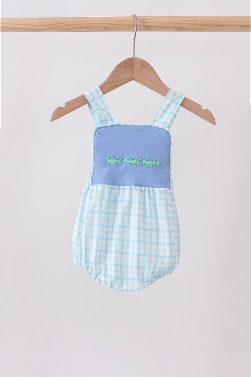 A baby romper in light blue and white gingham fabric with a solid light blue yoke panel featuring embroidered green letters, hanging on a wooden hanger against a plain light background.