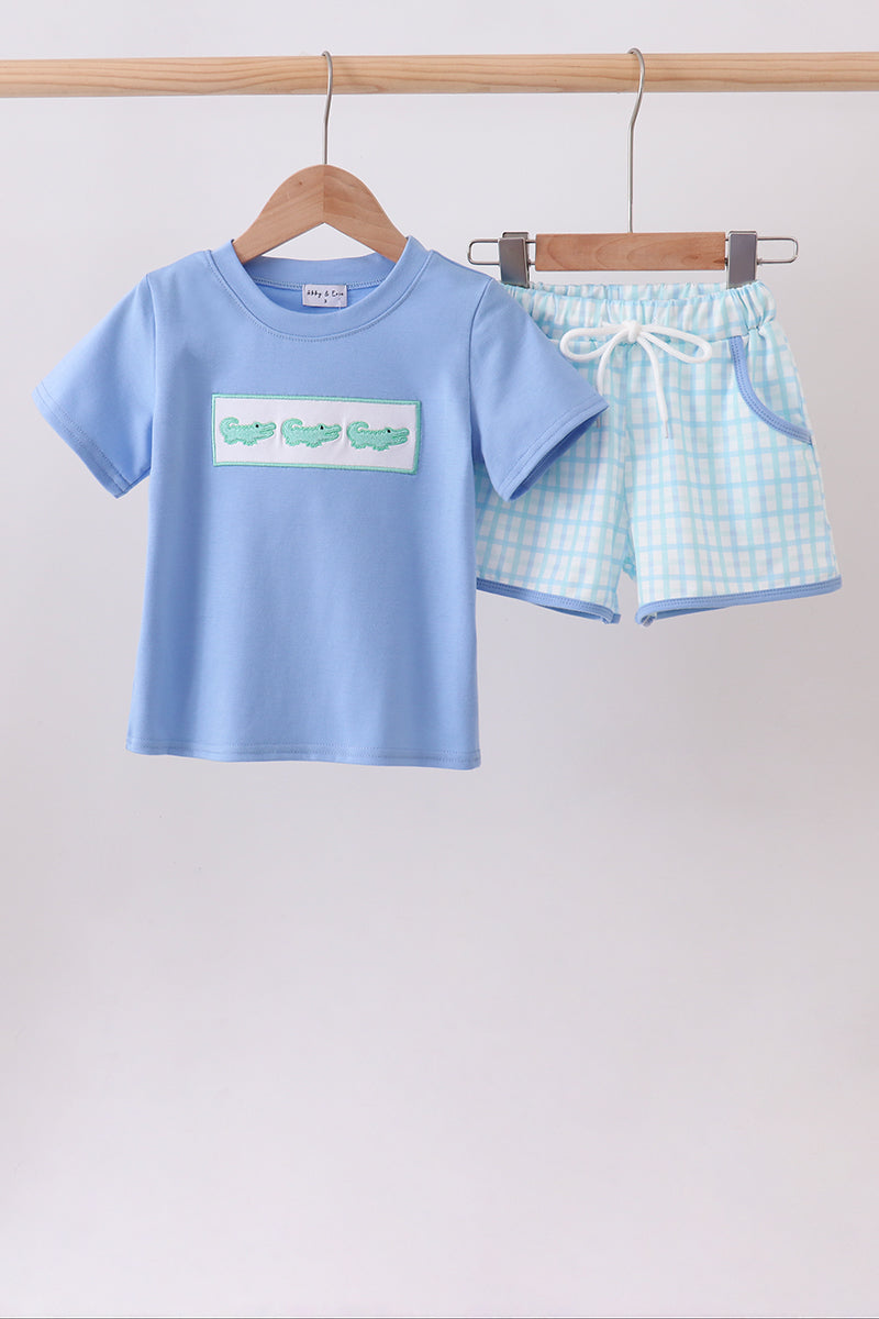 A two-piece children's outfit hanging on a wooden rod, featuring a light blue short-sleeve T-shirt with a green crocodile graphic on the chest and matching light blue and white checkered shorts with a white drawstring.