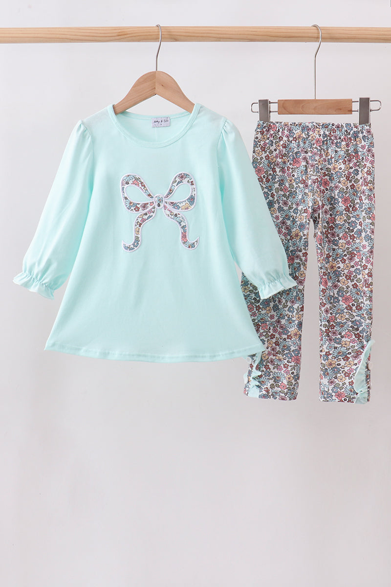 A two-piece children's outfit set hanging on wooden hangers against a plain light background. The set includes a light mint green long-sleeve top with a bow-shaped floral applique on the chest, and matching floral print pants in a multicolor pattern.