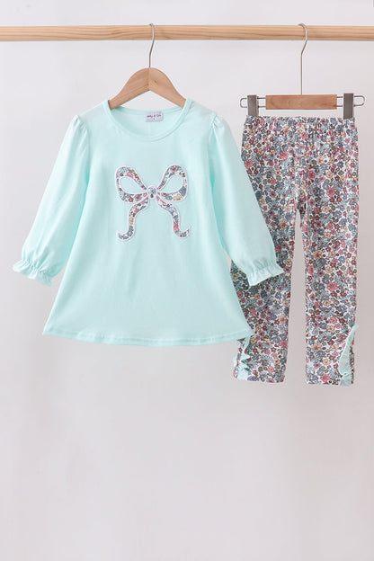 A two-piece children's outfit set hanging on wooden hangers against a plain light background. The set includes a light mint green long-sleeve top with a bow-shaped floral applique on the chest, and matching floral print pants in a multicolor pattern.