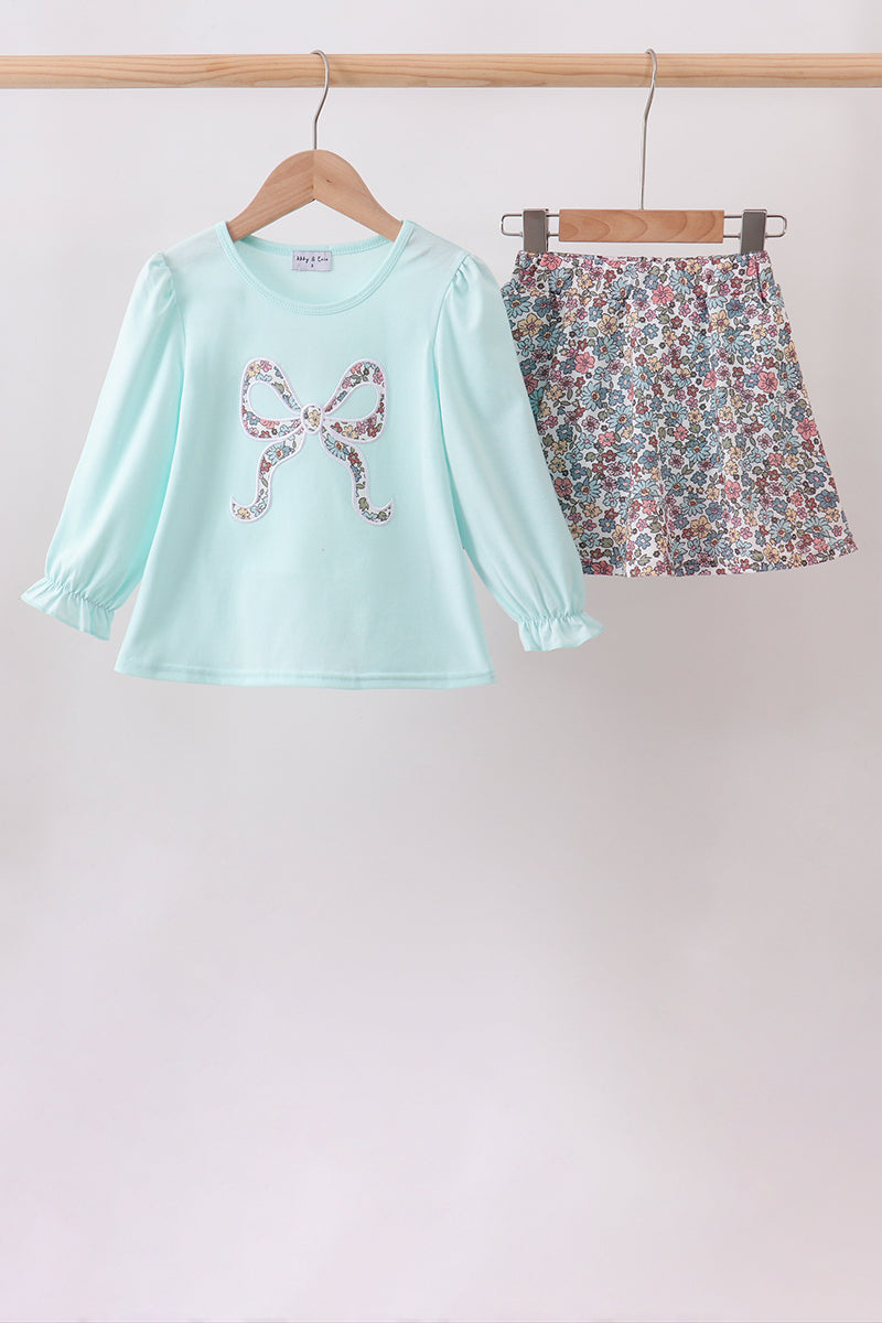 A two-piece girls' outfit hanging on wooden hangers from a wooden rod. The set includes a light mint green long-sleeve top with a sequined bow graphic on the chest, and a matching floral print mini skirt with pink, green, and blue flowers.