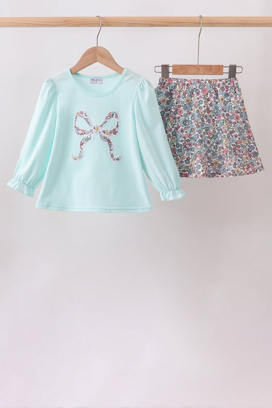 A two-piece girls' outfit hanging on wooden hangers from a wooden rod. The set includes a light mint green long-sleeve top with a sequined bow graphic on the chest, and a matching floral print mini skirt with pink, green, and blue flowers.