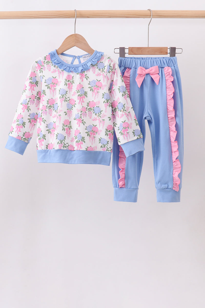 A two-piece children's outfit hanging on wooden hangers against a plain light background. The set includes a long-sleeve top with a white base, pink and blue floral print, and light blue ruffled trim at the collar, cuffs, and hem, paired with light blue pants featuring pink ruffle side panels, an elastic waistband, and a matching pink bow at the hips.
