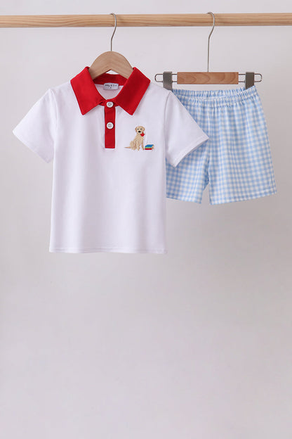 A children's two-piece outfit set hanging on wooden hangers against a plain light gray wall. The set includes a white short-sleeve polo shirt with a bright red collar and a small dog embroidery on the left chest, paired with light blue and white gingham check shorts.