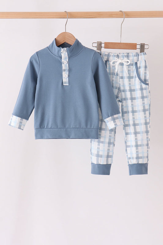 A two-piece baby clothing set hanging on wooden hangers from a wooden rod. The set includes a dusty blue long-sleeve mock-neck top with a white plaid collar and cuffs, and matching blue and white plaid pants with a white drawstring waist.