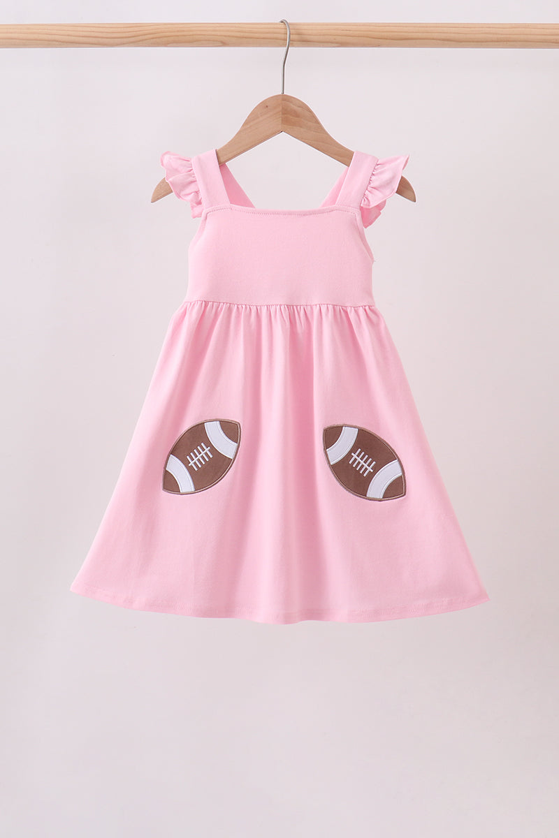 A pink sleeveless baby dress with ruffled shoulder straps and two football patches on the front, hanging on a wooden hanger against a light pink background.