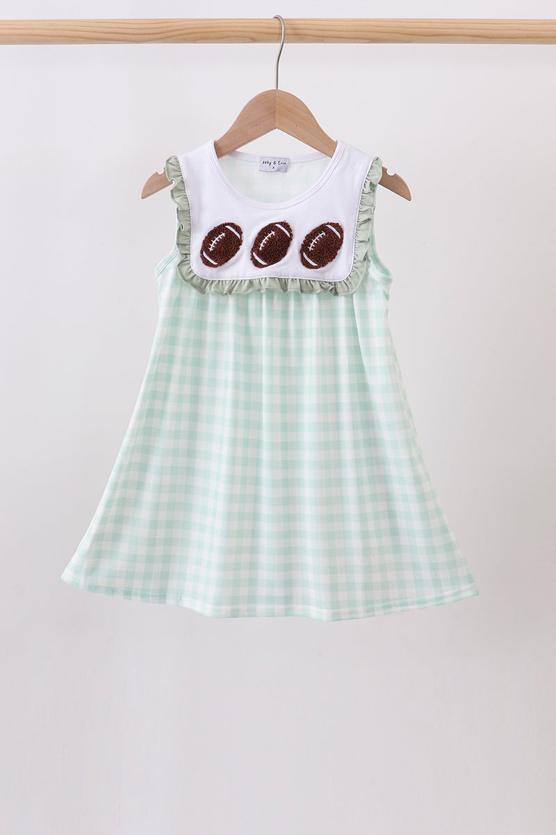 A sleeveless baby dress hanging on a wooden hanger against a plain light background. The dress has a white yoke with three brown football appliqués and ruffled trim, paired with a pale mint green and white gingham skirt.