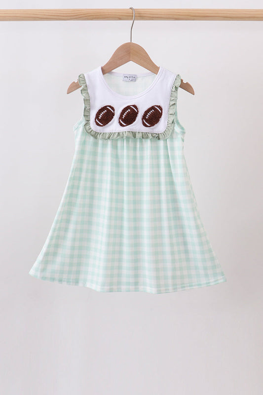 A sleeveless baby dress hanging on a wooden hanger against a plain light background. The dress has a white yoke with three brown football appliqués and ruffled trim, paired with a pale mint green and white gingham skirt.