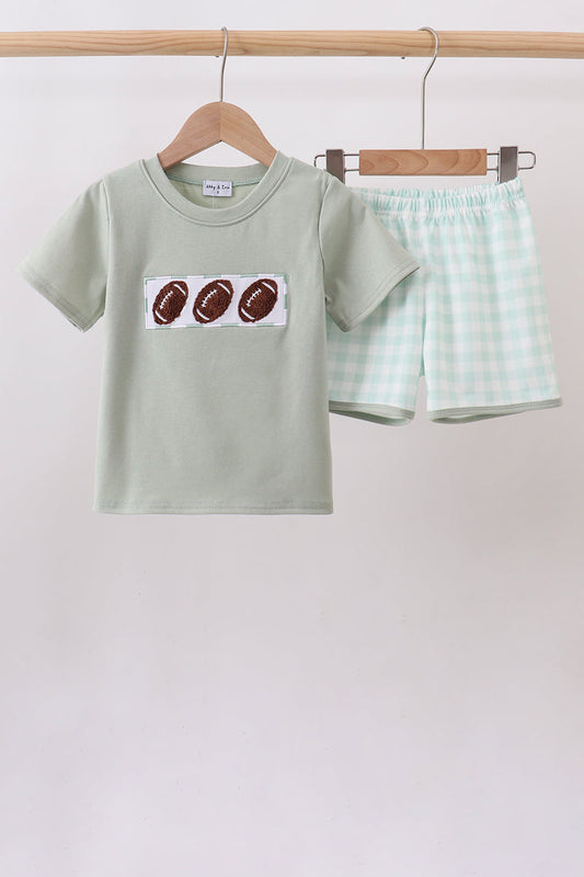 A children's two-piece outfit set hanging on a wooden hanger bar. The set includes a sage green short-sleeve t-shirt with a graphic of three brown footballs on the chest, and matching sage green and white gingham shorts.