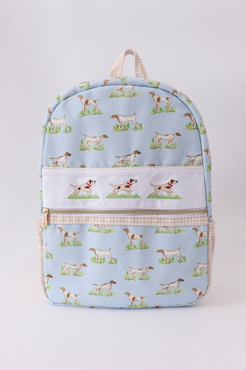 A light blue backpack featuring a repeating pattern of hunting dogs on grass, with a white front pocket and beige gingham trim, shown against a plain light gray background.