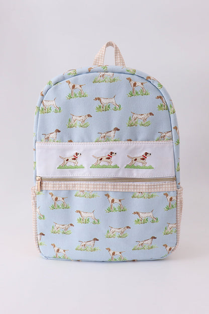 A light blue backpack featuring a repeating pattern of hunting dogs on grass, with a white front pocket and beige gingham trim, shown against a plain light gray background.