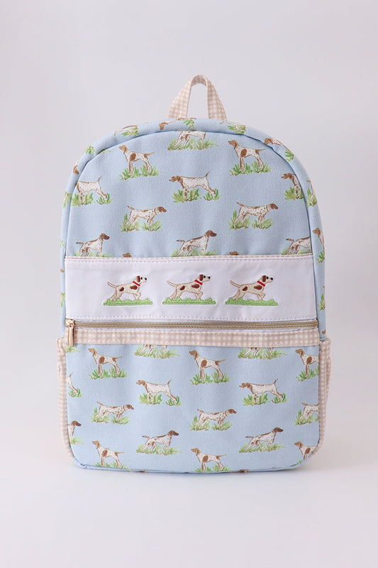 A light blue backpack featuring a repeating pattern of hunting dogs on grass, with a white front pocket and beige gingham trim, shown against a plain light gray background.