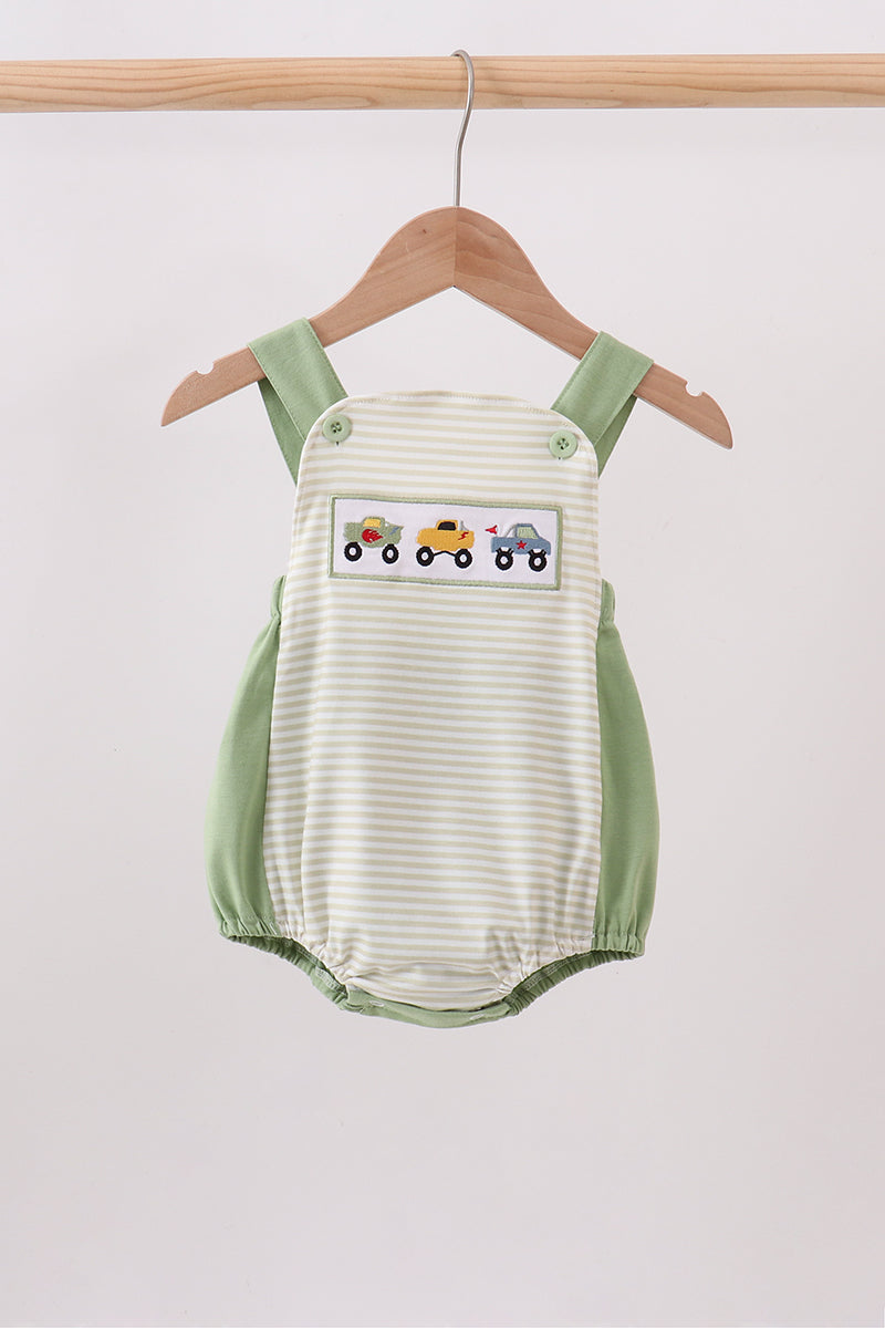 A baby romper with a cream and light green stripe pattern, featuring sage green shoulder straps and side panels, and a decorative patch of colorful toy trains on the chest. The romper is shown hanging on a wooden hanger from a wooden clothes rack against a plain light gray background.