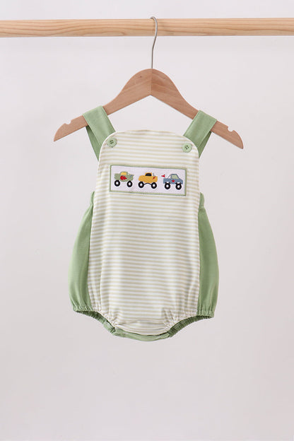 A baby romper with a cream and light green stripe pattern, featuring sage green shoulder straps and side panels, and a decorative patch of colorful toy trains on the chest. The romper is shown hanging on a wooden hanger from a wooden clothes rack against a plain light gray background.