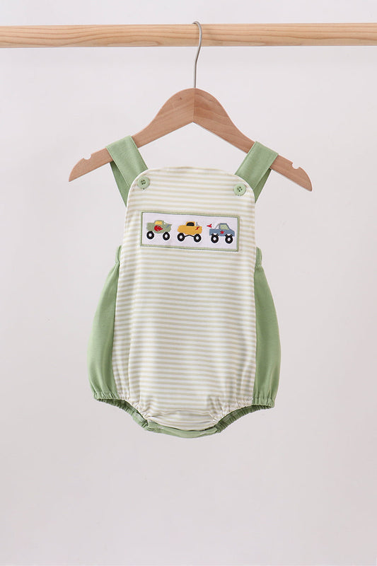 A baby romper with a cream and light green stripe pattern, featuring sage green shoulder straps and side panels, and a decorative patch of colorful toy trains on the chest. The romper is shown hanging on a wooden hanger from a wooden clothes rack against a plain light gray background.