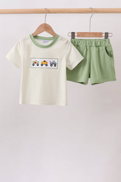 A children's two-piece outfit set consisting of a short-sleeve cream and green striped t-shirt with a colorful construction vehicle graphic on the chest, and matching sage green shorts, hanging on wooden hangers from a wooden clothing rack.