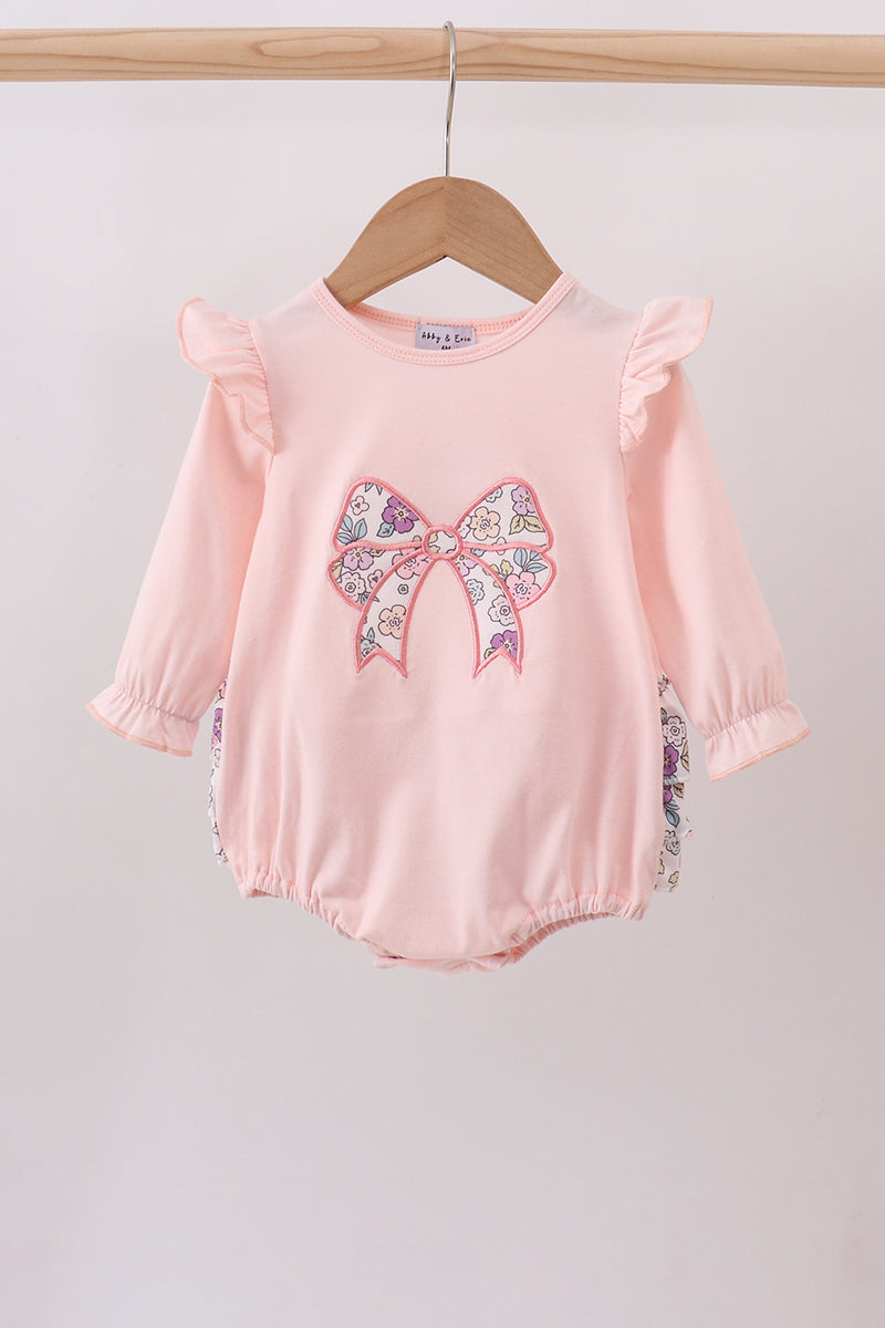 Pink baby romper with a large bow design on 
a wooden hanger against a light background