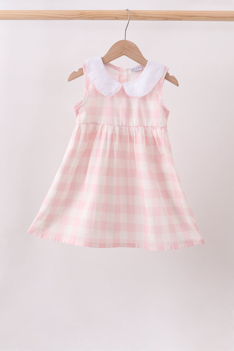 A sleeveless young girl's dress with a pink and white gingham pattern and a white Peter Pan collar, hanging on a wooden hanger from a light wood rod against a plain light background.