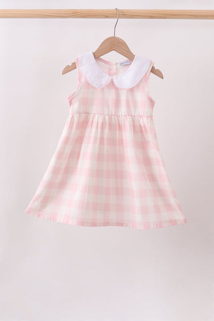 A sleeveless young girl's dress with a pink and white gingham pattern and a white Peter Pan collar, hanging on a wooden hanger from a light wood rod against a plain light background.