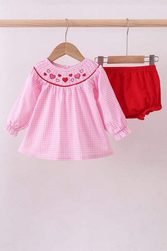 Pink checkered dress with heart details and red bloomers on hangers against a light background
