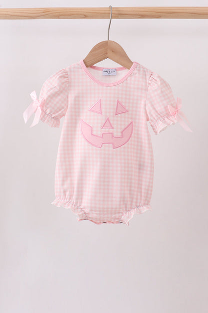 A baby girl's short-sleeve romper in a light pink and white gingham pattern, featuring a pink embroidered jack-o'-lantern face on the front and pink ribbon bows on the sleeves. The romper is hanging on a wooden hanger from a wooden clothes rack against a plain light gray background.