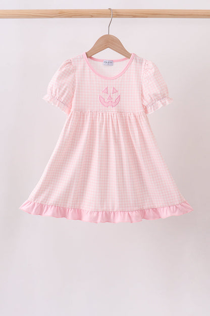 A light pink and white gingham baby dress with short flutter sleeves, a ruffled hem, and a pink embroidered pumpkin design on the chest, hanging on a wooden hanger against a light background.