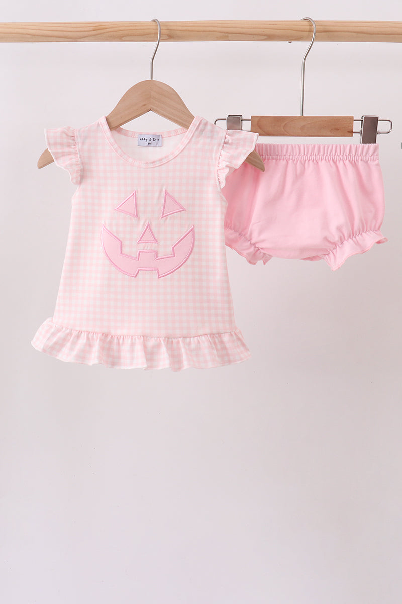 A baby girl's two-piece Halloween outfit consisting of a light pink and white gingham short-sleeve top with a pink pumpkin face applique, and matching pink ruffled bloomers, hanging on wooden hangers from a wooden rod against a plain light background.