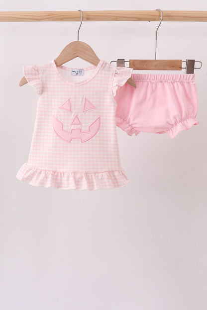 A baby girl's two-piece Halloween outfit consisting of a light pink and white gingham short-sleeve top with a pink pumpkin face applique, and matching pink ruffled bloomers, hanging on wooden hangers from a wooden rod against a plain light background.