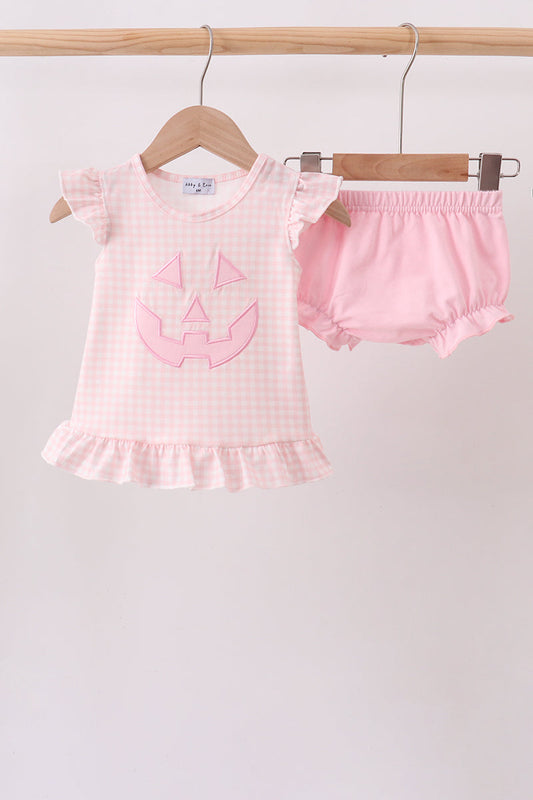 A baby girl's two-piece Halloween outfit consisting of a light pink and white gingham short-sleeve top with a pink pumpkin face applique, and matching pink ruffled bloomers, hanging on wooden hangers from a wooden rod against a plain light background.