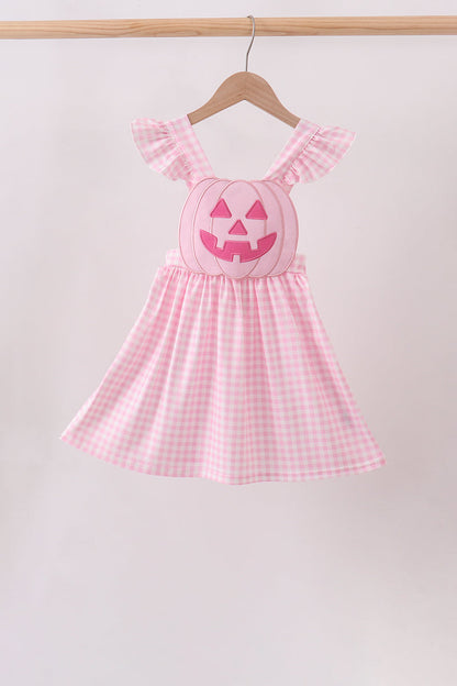 A pink and white gingham baby dress with ruffled shoulder straps and a smocked pumpkin face on the chest, hanging on a wooden hanger from a wooden rod against a light background.