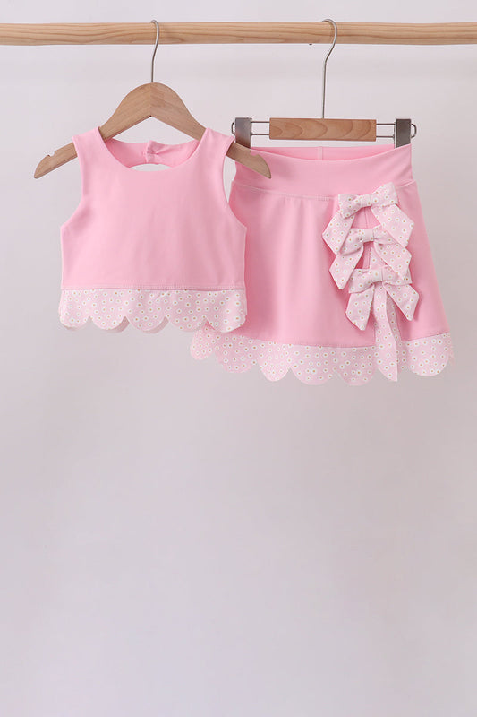A two-piece girls' outfit set in light pink, hanging on wooden hangers from a wooden rod. The set includes a sleeveless top with a scalloped eyelet lace hem and matching shorts featuring a large ruffled bow on the left side and a matching scalloped eyelet lace hem.
