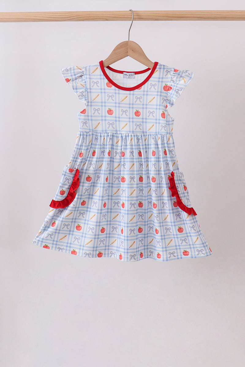 A toddler girl's short-sleeve dress with a white and light blue gingham pattern, decorated with red apple motifs and red ruffle trim, hanging on a wooden hanger against a plain light gray wall.