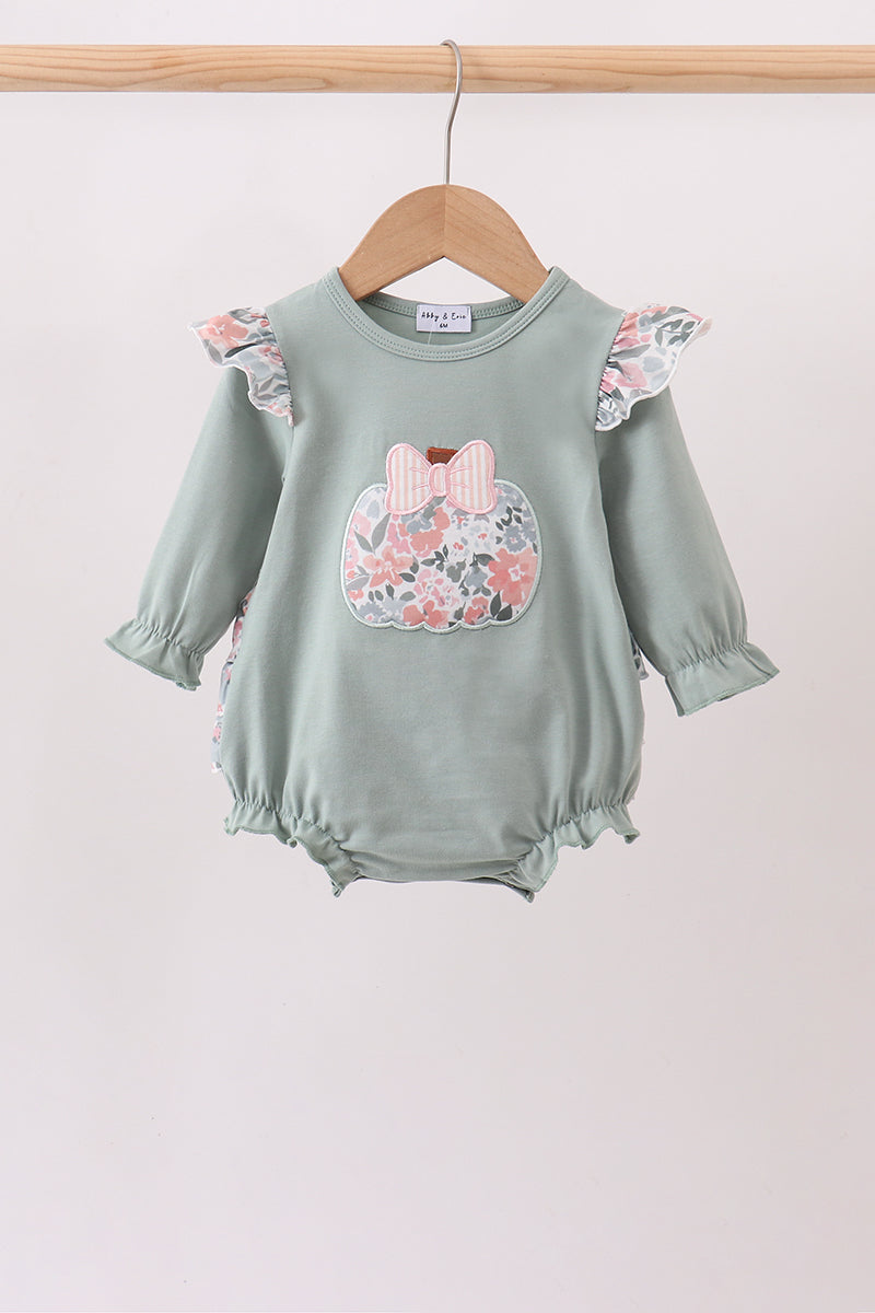 A baby long-sleeve romper in sage green, hanging on a wooden hanger. The romper features ruffled sleeves and hem, a floral applique chest design with a pink bow, and ruffled trim along the side seams.