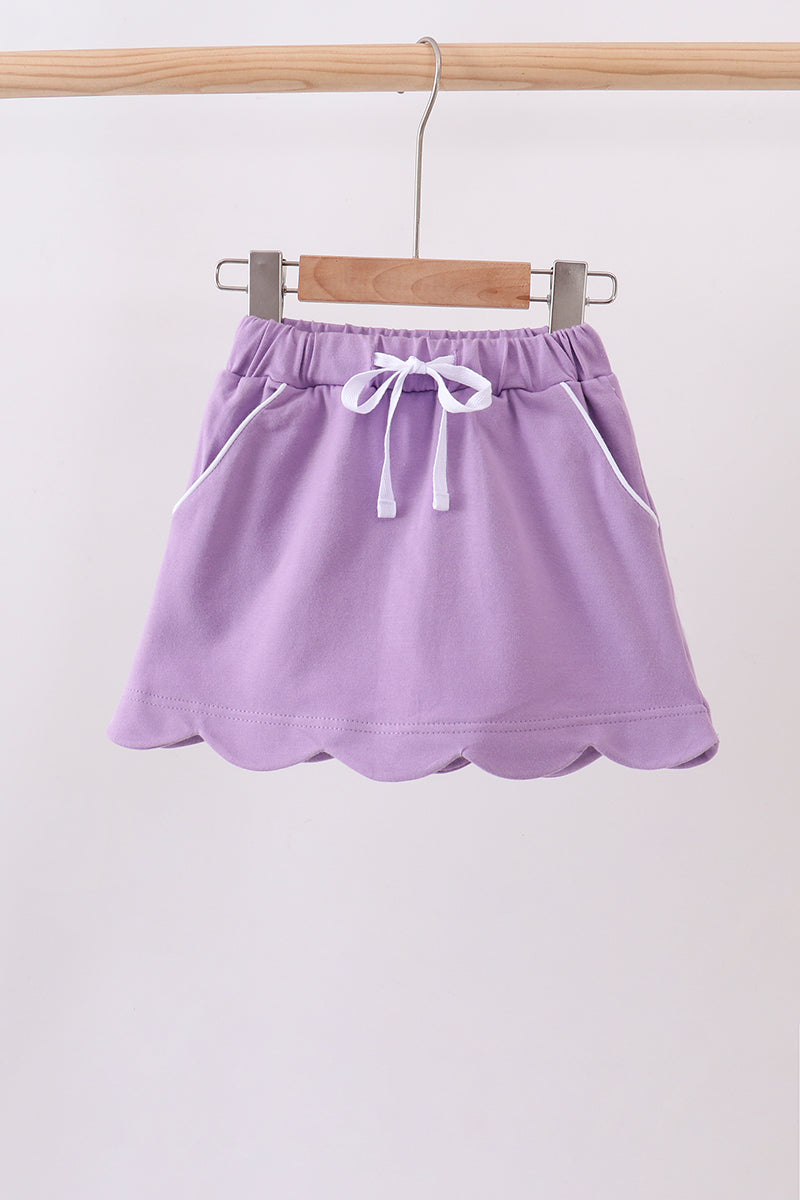 A lavender purple mini skirt with a scalloped hem, elastic waistband, and white drawstring, hanging from a wooden hanger on a clothing rack.