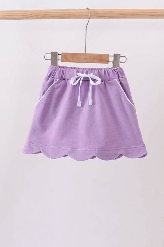 A lavender purple mini skirt with a scalloped hem, elastic waistband, and white drawstring, hanging from a wooden hanger on a clothing rack.