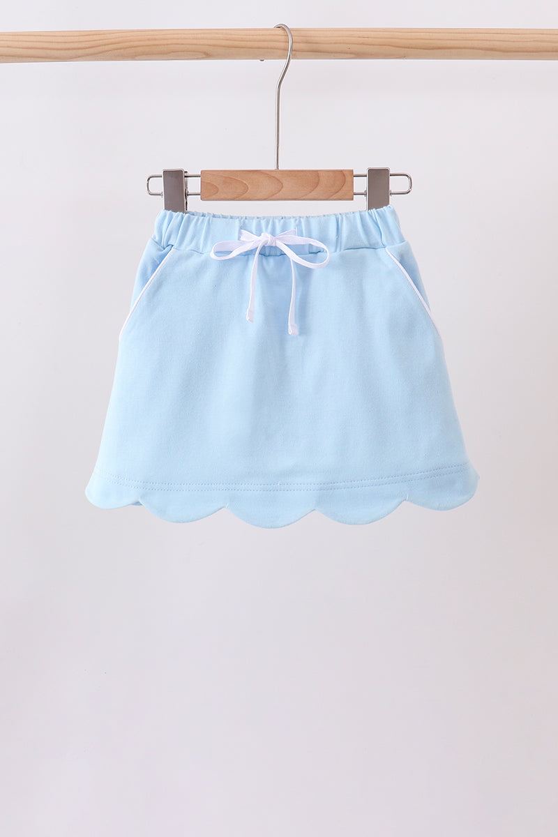 A light blue cotton skirt with a scalloped hem and a white drawstring, hanging on a wooden hanger from a wooden clothes rack against a plain light background.