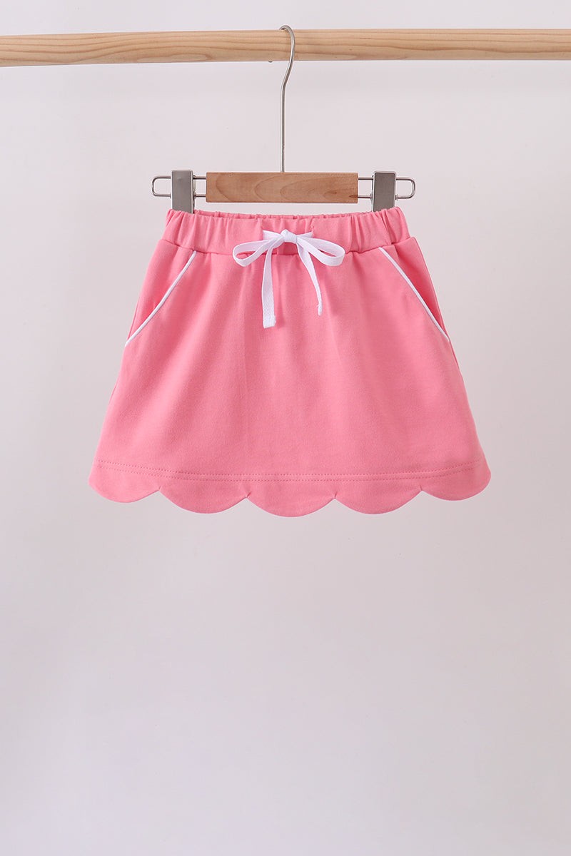 A pink mini skirt with a scalloped hem, white drawstring, and white piping details, hanging on a wooden hanger from a wooden clothes rack.