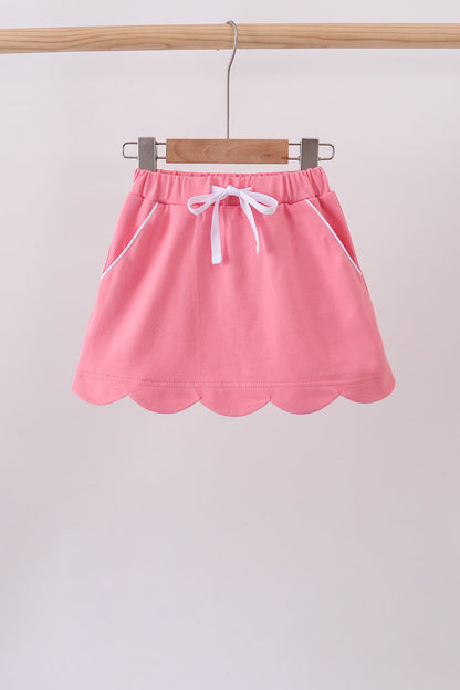 A pink mini skirt with a scalloped hem, white drawstring, and white piping details, hanging on a wooden hanger from a wooden clothes rack.