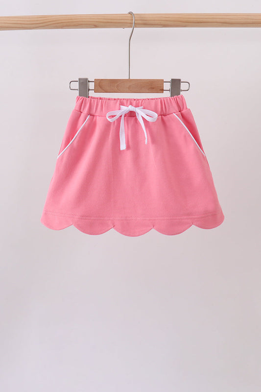 A pink mini skirt with a scalloped hem, white drawstring, and white piping details, hanging on a wooden hanger from a wooden clothes rack.
