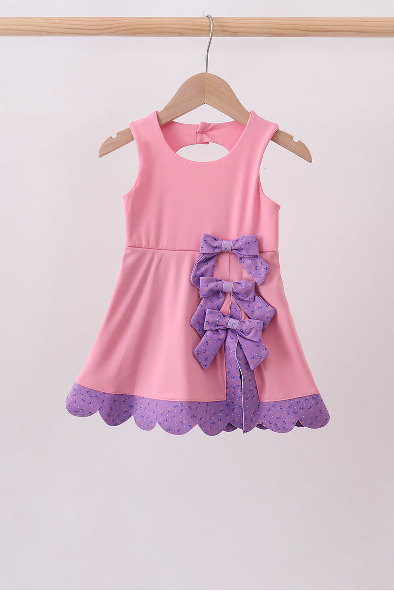 A pink sleeveless girls' dress hanging on a wooden hanger against a plain light background. The dress has a large purple ruffled bow at the center front and a purple scalloped hem with a subtle polka dot pattern.