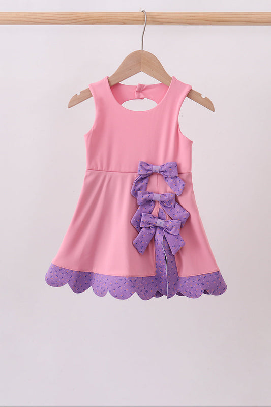 A pink sleeveless girls' dress hanging on a wooden hanger against a plain light background. The dress has a large purple ruffled bow at the center front and a purple scalloped hem with a subtle polka dot pattern.