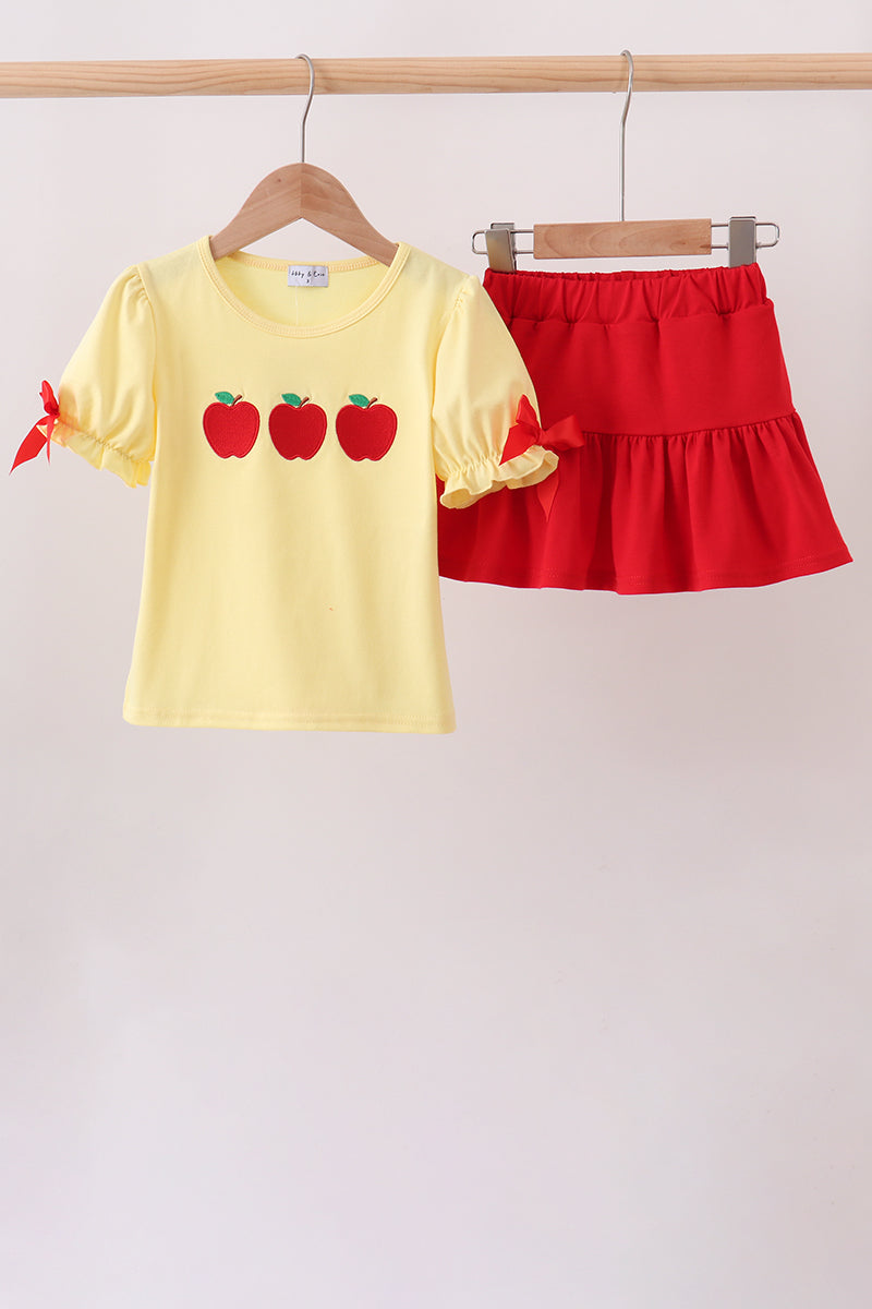 A two-piece children's outfit hanging on wooden hangers from a wooden rod. The set includes a pale yellow short-sleeve top with three red apple graphics on the chest and red ribbon bows on the sleeves, paired with a bright red tiered ruffle skirt.