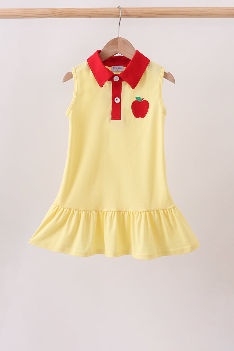 A sleeveless yellow children's dress hanging on a wooden hanger against a light beige wall. The dress has a red collar and red button placket, with a red apple applique on the left chest.