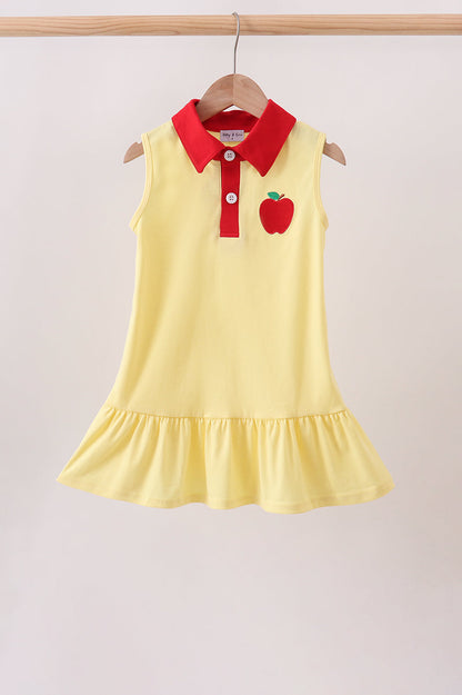A sleeveless yellow children's dress hanging on a wooden hanger against a light beige wall. The dress has a red collar and red button placket, with a red apple applique on the left chest.