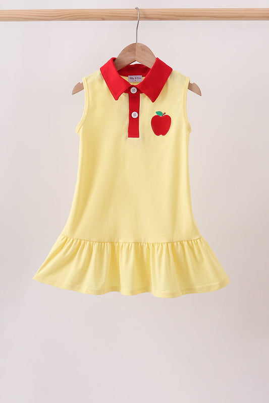 A sleeveless yellow children's dress hanging on a wooden hanger against a light beige wall. The dress has a red collar and red button placket, with a red apple applique on the left chest.